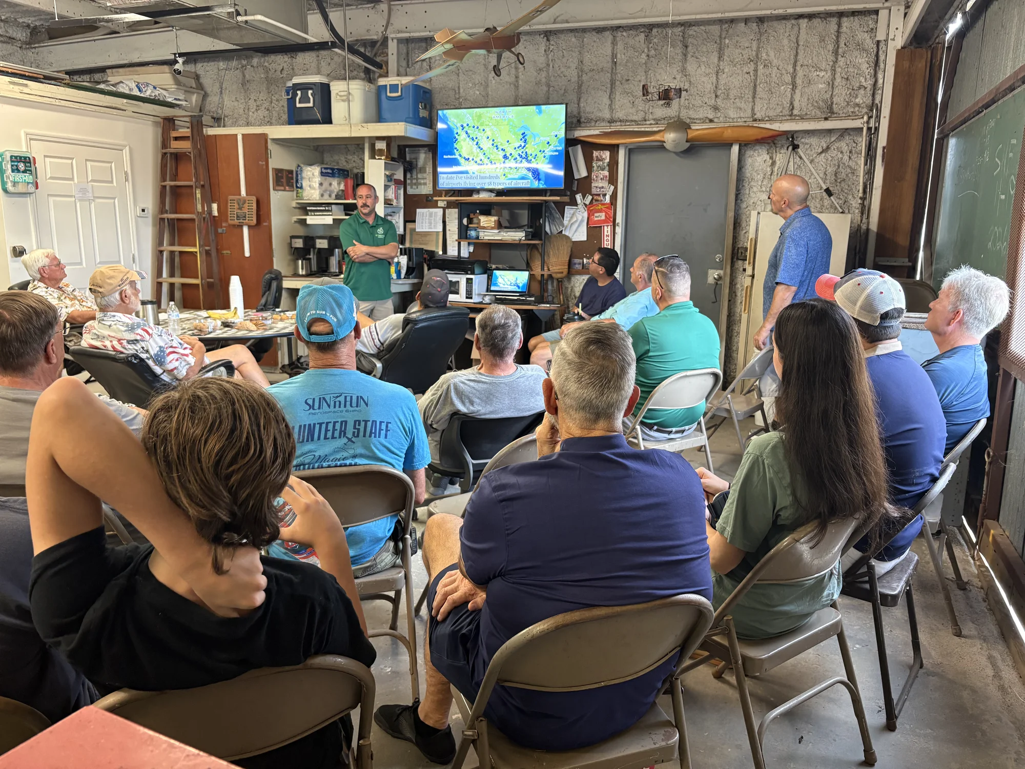 Chapter members in folding chairs facing the TV at Hangar 100 during Patrick Blackwelder's presentation